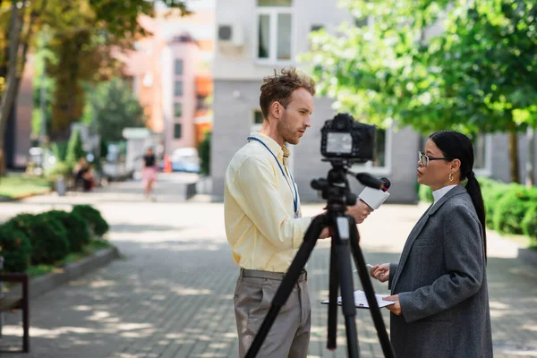 Side view of newscaster holding microphone near asian businesswoman in glasses on street — Stock Photo