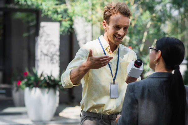 Happy reporter gesturing while taking interview with blurred asian businesswoman in glasses — Stock Photo