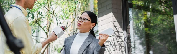 Reporter with microphone making reportage with asian businesswoman in glasses holding cellphone, banner — Stock Photo