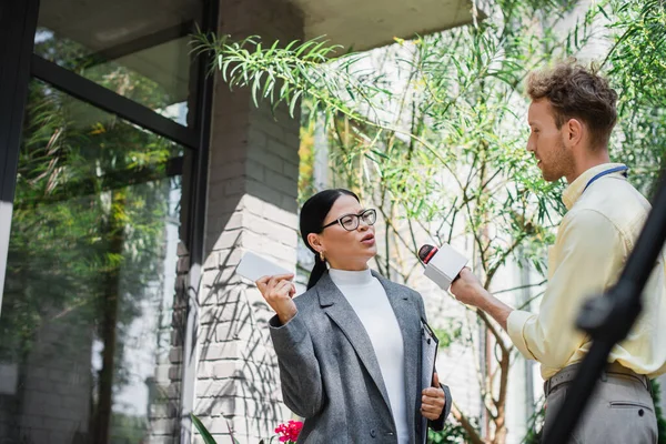 Reporter with microphone making reportage with asian businesswoman in glasses holding cellphone — Stock Photo