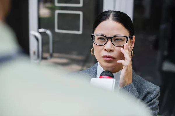 Borrosa reportera con micrófono cerca pensativo asiático mujer de negocios ajuste gafas - foto de stock