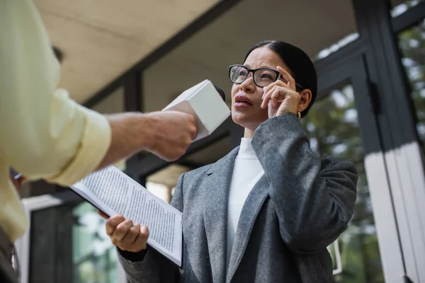 Bajo ángulo vista de reportero con micrófono cerca pensativo asiático mujer de negocios ajuste gafas - foto de stock