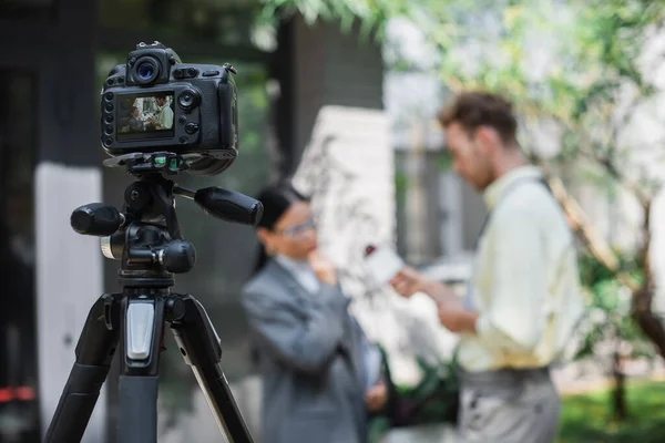 Câmera digital com repórter segurando microfone ao tomar entrevista de empresária asiática em óculos na tela — Fotografia de Stock