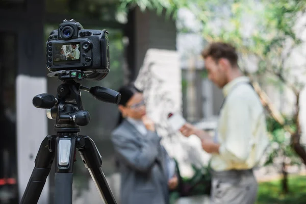 Digitalkamera mit Reporter, der Mikrofon hält, während er ein Interview mit asiatischer Geschäftsfrau mit Brille auf dem Bildschirm führt — Stockfoto