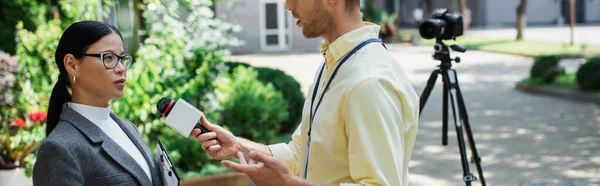 Journalist gesturing while holding microphone and taking interview of asian businesswoman in glasses, banner — Stock Photo