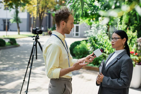 Journalistin gestikuliert, während sie das Mikrofon in der Hand hält und ein Interview mit einer asiatischen Geschäftsfrau mit Brille führt — Stockfoto