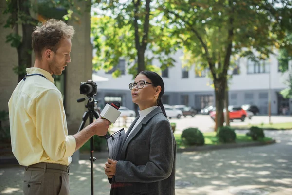 Journalist holding microphone and taking interview of asian businesswoman in glasses near digital camera on street — Stock Photo