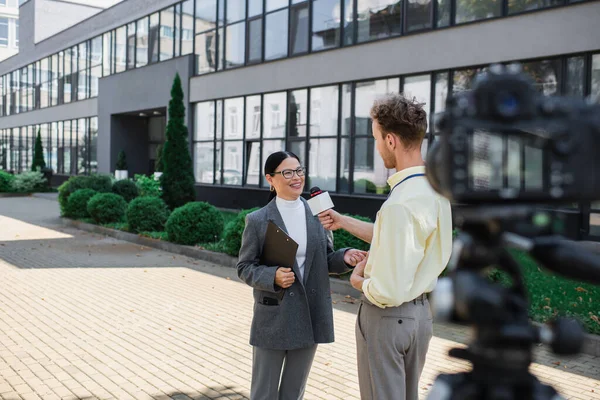Journalist holding microphone and taking interview of cheerful asian businesswoman in glasses standing near building — Stock Photo