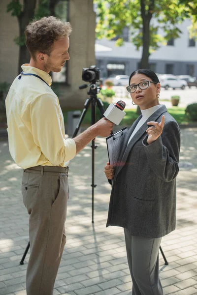 Periodista sosteniendo micrófono y tomando entrevista de asiático mujer de negocios en gafas sosteniendo portapapeles y apuntando lejos - foto de stock