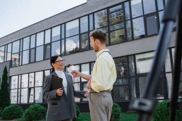 Nachrichtensprecher hält Mikrofon in der Hand und interviewt fröhliche asiatische Geschäftsfrau in Brille, die in der Nähe des Gebäudes steht — Stockfoto