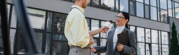 Nachrichtensprecher hält Mikrofon in der Hand und interviewt fröhliche asiatische Geschäftsfrau in Brille, die in der Nähe des Gebäudes steht, Banner — Stockfoto