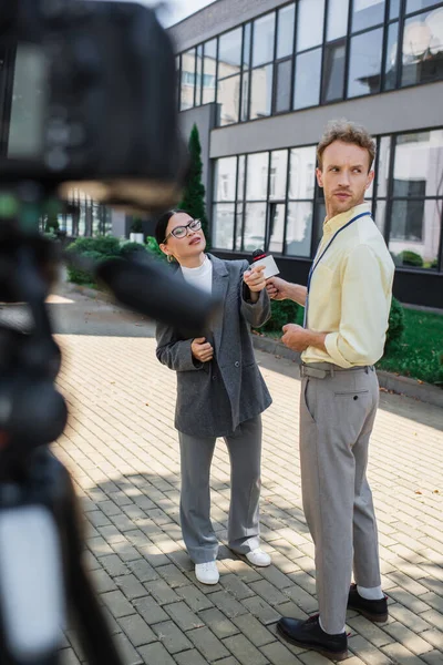 Asian businesswoman in glasses holding clipboard and pointing away near distracted journalist with microphone and blurred digital camera — Stock Photo