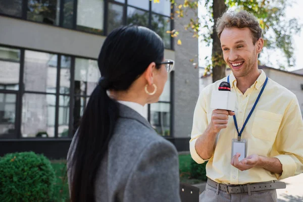 Happy journalist smiling while holding microphone and looking at blurred asian businesswoman — Stock Photo