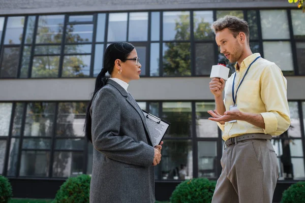 Asiatische Geschäftsfrau in Brille mit Klemmbrett und Blick auf Journalist mit Mikrofon — Stockfoto