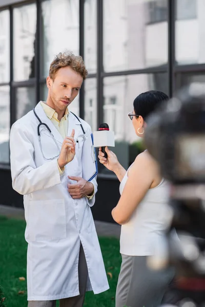 Arzt in weißem Mantel zeigt mit Finger und Warnung, während er einem asiatischen Journalisten mit Mikrofon ein Interview gibt — Stockfoto