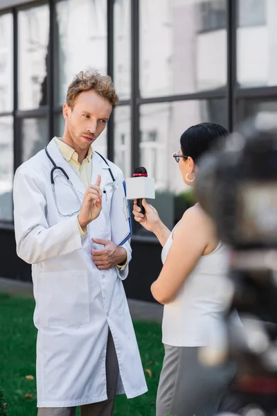Médecin en manteau blanc pointant du doigt et avertissement tout en donnant une interview à un journaliste asiatique avec microphone — Photo de stock