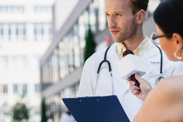 Blurred journalist holding microphone near doctor in white coat — Stock Photo