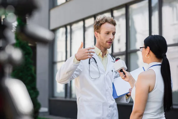 Docteur en manteau blanc tenant presse-papiers et geste tout en donnant une interview à un journaliste asiatique avec microphone — Photo de stock