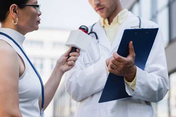 Брюнетка азіатський журналіст тримає мікрофон біля лікаря в білому пальто з буфером — стокове фото