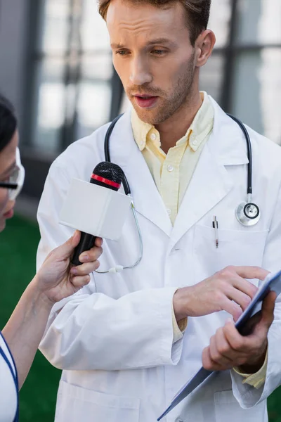 Docteur en manteau blanc tenant presse-papiers et gestuelle tout en donnant une interview à floue journaliste asiatique avec microphone — Photo de stock