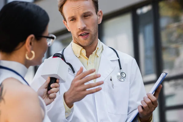 Doctor in white coat holding clipboard and gesturing while giving interview to blurred and tattooed asian journalist with microphone — Stock Photo