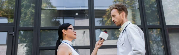 Vista laterale del giornalista asiatico che tiene il microfono mentre prende l'intervista del medico in cappotto bianco, banner — Foto stock