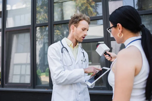 Doctor in white coat pointing at digital tablet while giving interview to blurred asian journalist with microphone — Stock Photo