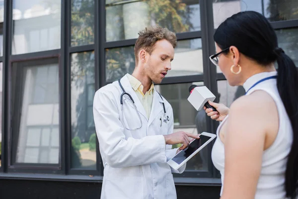 Médecin en manteau blanc pointant vers tablette numérique tout en donnant une interview à floue journaliste asiatique avec microphone — Photo de stock
