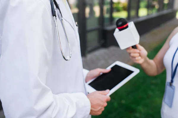 Vue recadrée du médecin en manteau blanc tenant tablette numérique tout en donnant une interview au journaliste avec microphone — Photo de stock