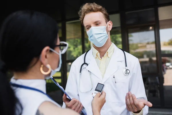Verschwommener asiatischer Reporter mit Stimmenrekorder in der Nähe eines Arztes in medizinischer Maske und weißem Mantel — Stockfoto
