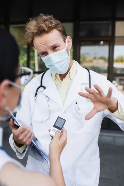 Размытый азиатский репортер держит диктофон рядом с доктором в медицинской маске и белом халате жестикулируя во время интервью — стоковое фото