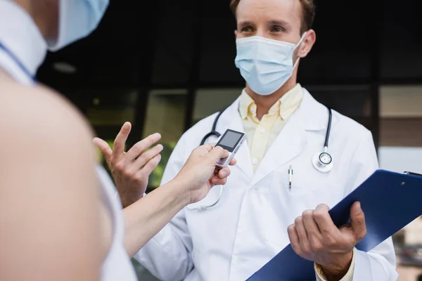 Verschwommener Reporter mit Stimmenrekorder in der Nähe von Arzt in medizinischer Maske und weißem Mantel gestikuliert während Interview — Stockfoto