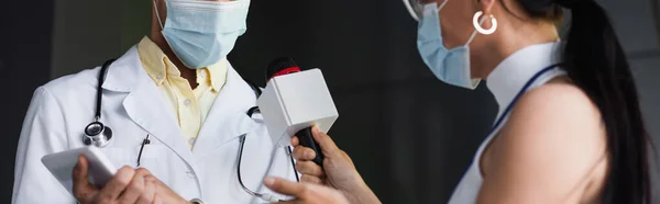 Vista recortada do repórter na máscara médica que prende o microfone perto do doutor no revestimento branco com tabuleta digital, bandeira — Fotografia de Stock