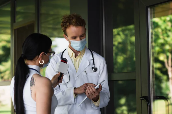Лікар в медичній масці вказує на цифровий планшет біля татуйованого журналіста з мікрофоном — стокове фото