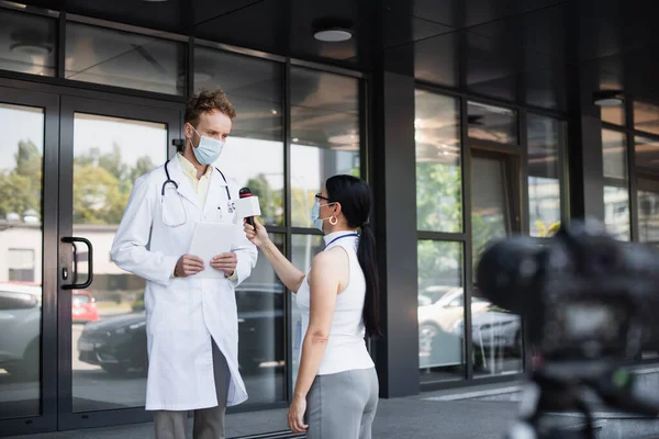 Азіатський репортер в медичній масці тримає мікрофон біля лікаря в білому пальто з цифровим планшетом, що стоїть біля клініки — стокове фото