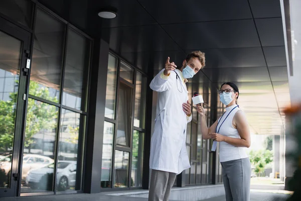 Arzt in weißem Mantel und medizinischer Maske zeigt weg, während er einem asiatischen Journalisten mit Mikrofon ein Interview gibt — Stockfoto