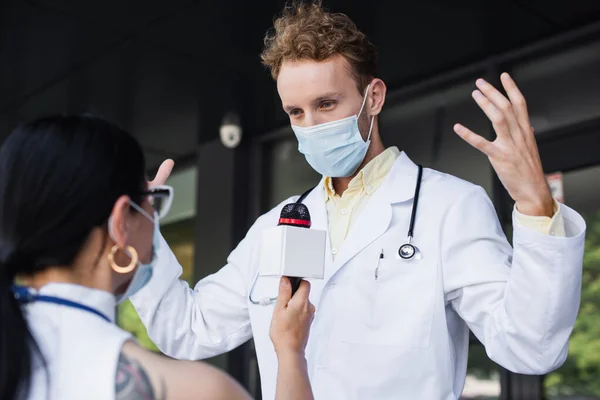Врач в медицинской маске жестикулирует во время интервью с размытым азиатским репортером — стоковое фото