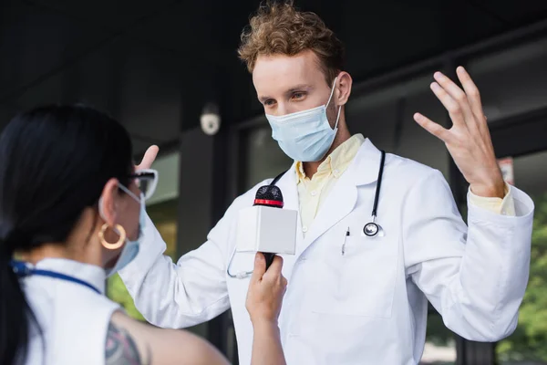 Médecin en masque médical gestuelle lors d'une interview avec flou asiatique journaliste — Photo de stock