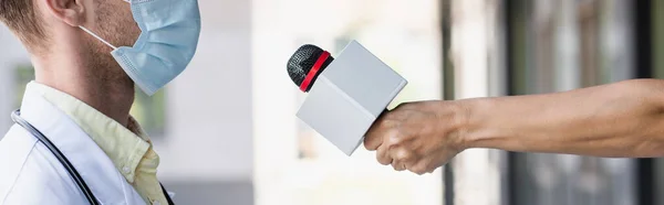 Обрізаний вид репортера, що тримає мікрофон біля лікаря в медичній масці, банер — стокове фото