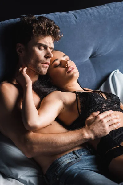 Sensuale uomo toccare bella donna in lingerie sul letto isolato su nero — Foto stock