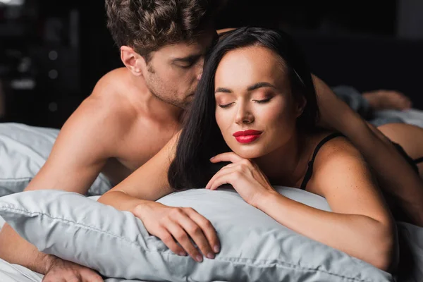 Hombre sin camisa abrazando a mujer sensual con labios rojos en la cama sobre fondo negro - foto de stock