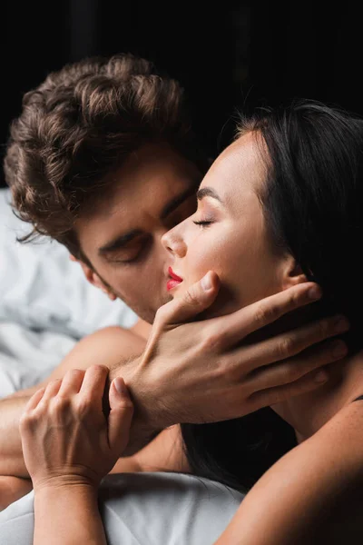 Man kissing and touching sexy brunette girlfriend on bed isolated on black — Stock Photo