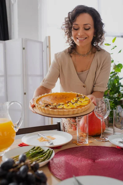 Усміхнена афро-американська жінка тримає смачний пиріг біля прикрашеного обіду з подяки. — стокове фото