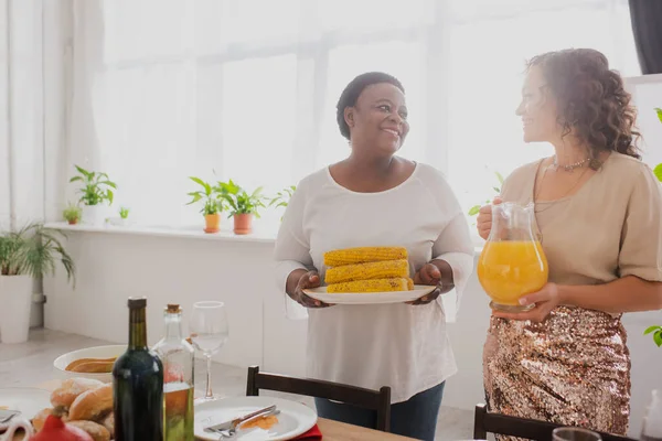 Mujer afroamericana sonriente sosteniendo jugo de naranja cerca de la madre con maíz y cena de acción de gracias - foto de stock