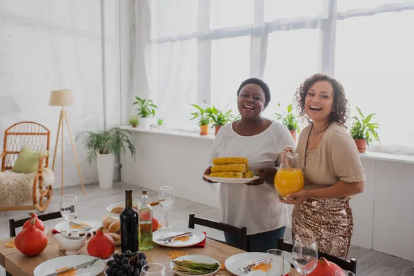 Mulheres americanas africanas felizes segurando milho e suco de laranja perto do jantar de ação de graças decorado — Fotografia de Stock