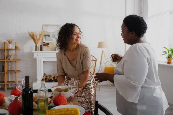 Улыбающаяся африканская американка смотрит на мать с апельсиновым соком рядом с праздничным ужином — стоковое фото