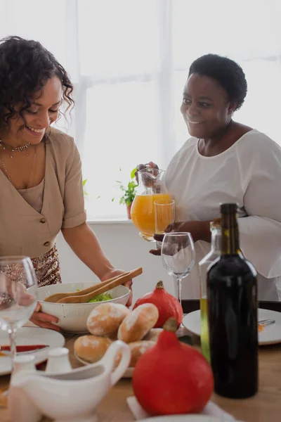 Mujer afroamericana sonriente con jugo de naranja mirando a su hija sirviendo la cena de Acción de Gracias - foto de stock