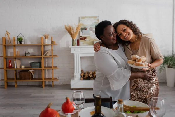 Happy african american woman holding buns and hugging mother near thanksgiving dinner — Stock Photo