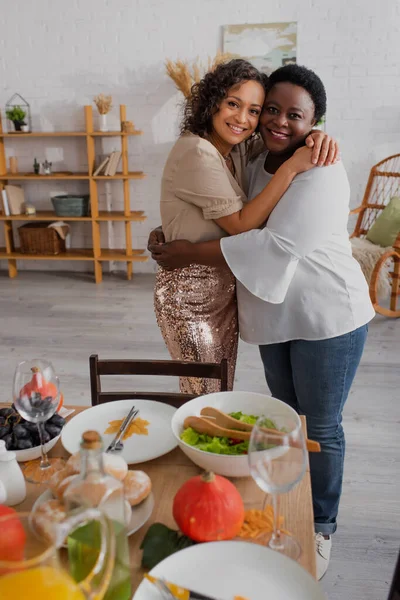 Mère et fille afro-américaine souriantes serrant dans leurs bras près du dîner d'Action de grâces flou à la maison — Photo de stock