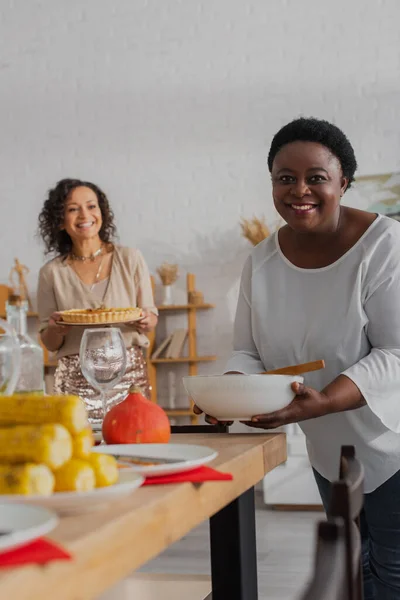 Madura afroamericana mujer sosteniendo plato cerca de la cena festiva y borrosa hija - foto de stock